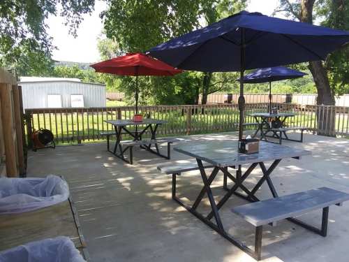 Outdoor seating area with picnic tables and umbrellas, surrounded by greenery and a wooden fence.