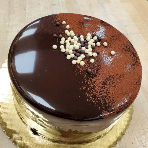 A glossy chocolate cake topped with decorative sprinkles and cocoa powder on a gold cake board.