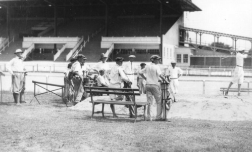 A historic black-and-white photo of a group of men at a baseball field, some seated on a bench, watching a game.