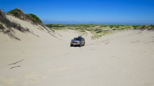 A vehicle driving through a sandy dune landscape under a clear blue sky.