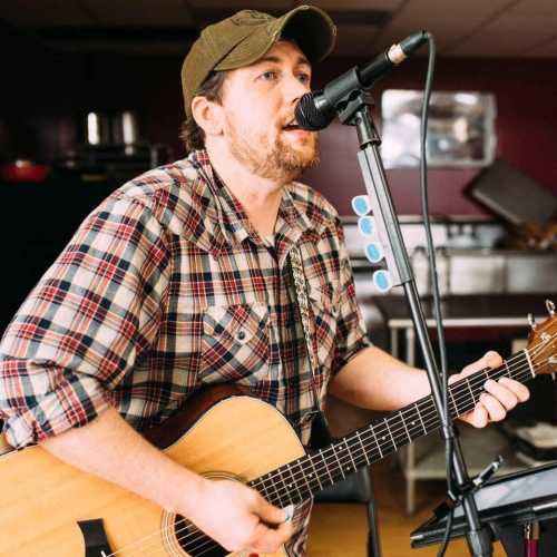 A musician in a plaid shirt sings into a microphone while playing an acoustic guitar.