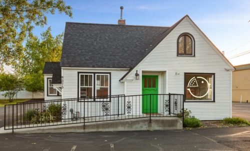 A charming white house with a green door, sloped roof, and a large window featuring a playful design.