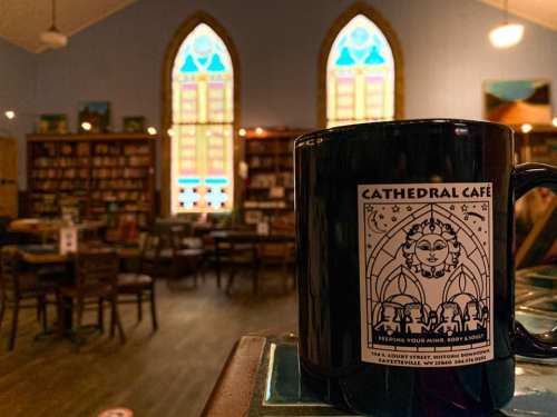 A coffee mug with "Cathedral Café" design in a cozy café with stained glass windows and wooden furniture.