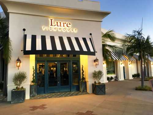 Exterior of Lure Fishhouse restaurant with a striped awning, palm trees, and evening lighting.