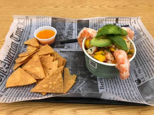 A bowl of shrimp with avocado and mango salsa, served with tortilla chips and a side of sauce on a newspaper-lined tray.