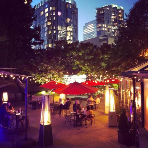 Outdoor dining area with red umbrellas, heaters, and city buildings in the background, illuminated at dusk.