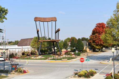 A giant wooden chair sculpture stands in a park, surrounded by trees and a clear blue sky.
