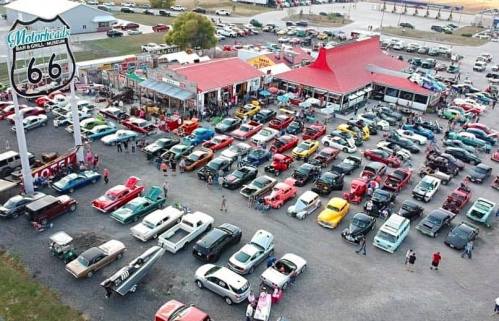 Aerial view of a bustling car show with numerous vintage cars parked around a diner and Route 66 sign.
