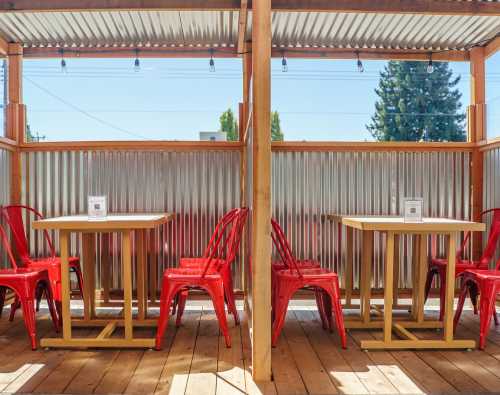 Two wooden tables with red chairs in a bright outdoor dining area, surrounded by corrugated metal walls.