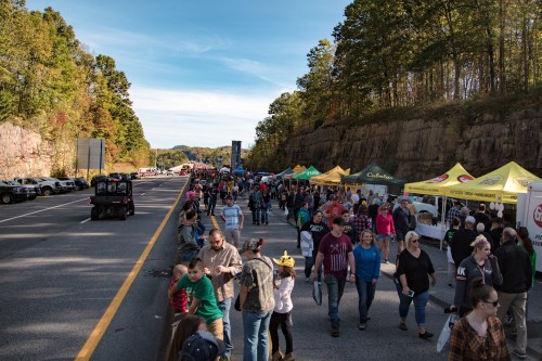 A busy outdoor event along a highway, with crowds, vendor tents, and autumn foliage in the background.