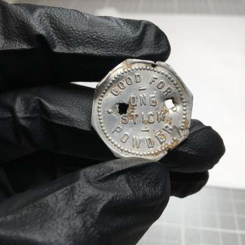A person in black gloves holds an old, worn token with the inscription "GOOD FOR ONE STICK POWDER" and two holes.