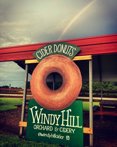 Sign for Windy Hill Orchard & Cidery featuring a large cider donut and a rainbow in the background.