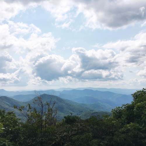 A scenic view of rolling mountains under a partly cloudy sky, with lush greenery in the foreground.
