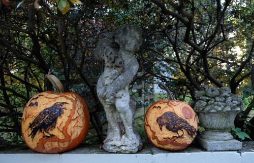 Two intricately carved pumpkins featuring crows, flanking a stone statue amidst lush greenery.
