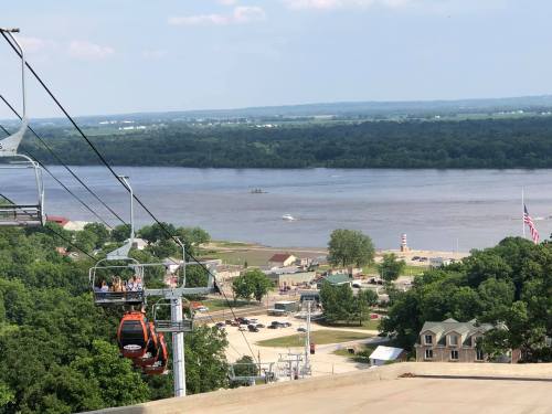 A scenic view from a gondola, overlooking a river, beach area, and green landscape under a clear blue sky.
