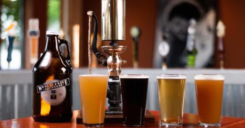 A growler and four glasses of beer in various colors on a wooden table, with taps in the background.