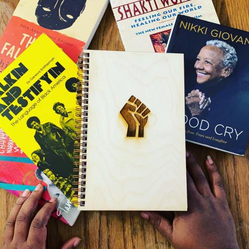 A wooden notebook with a raised fist design, surrounded by books on Black culture and literature. Hands are holding the notebook.