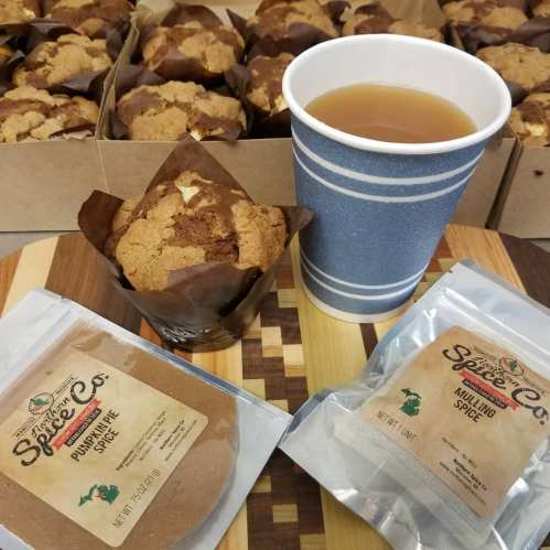 A box of pumpkin spice muffins, a cup of tea, and packets of pumpkin and mulling spices on a wooden surface.