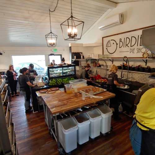 A busy pie shop kitchen with staff preparing food, serving customers, and a display case filled with pies.
