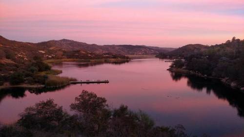 Serene lake at sunset, surrounded by hills and trees, with soft pink and purple hues in the sky reflecting on the water.