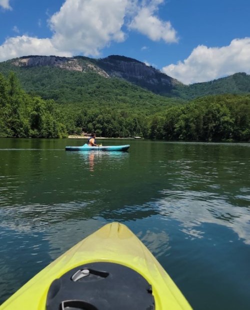 A person kayaking on a calm lake surrounded by lush green mountains and a blue sky with fluffy clouds.