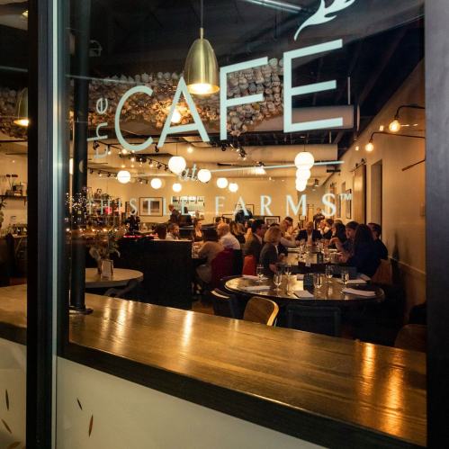 Interior of a cafe with diners at tables, warm lighting, and a sign reading "the CAFE at the FARM."