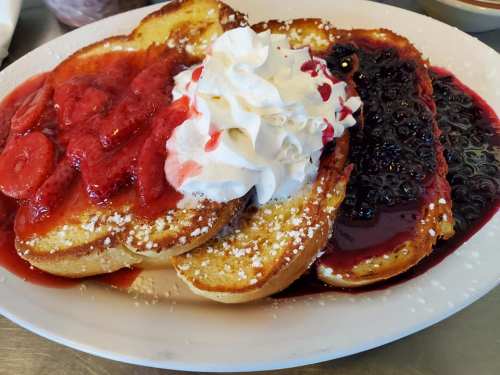 Three slices of French toast topped with strawberry and blackberry sauces, whipped cream, and powdered sugar.
