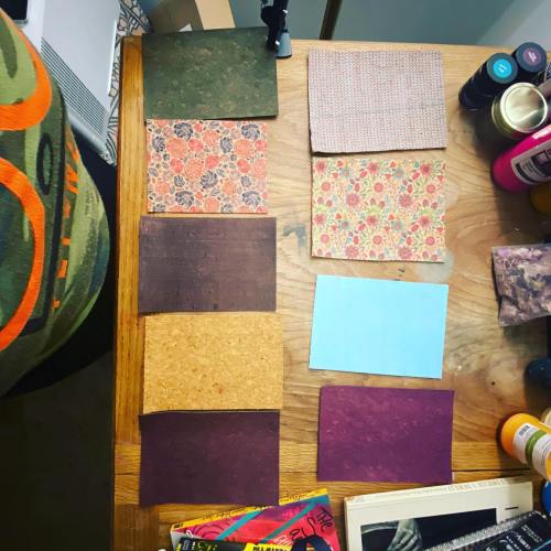 A wooden table displays various textured paper samples in different colors and patterns, alongside art supplies.