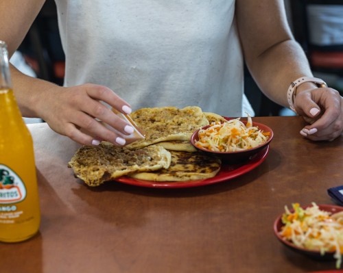A person sprinkles seasoning on a plate of flatbreads and coleslaw, with a bottle of orange soda nearby.