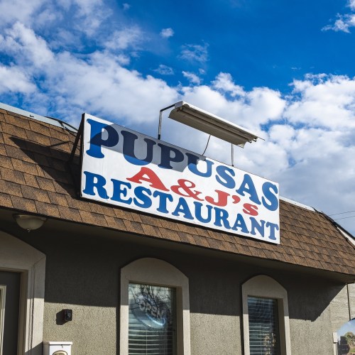 Sign for "Pupusas A & J's Restaurant" on a building with a blue sky and clouds in the background.