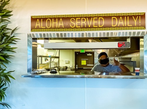 A person in a mask smiles and gives a thumbs up from behind a food service counter with a sign that says "Aloha Served Daily!"