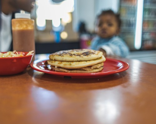 A stack of pancakes on a red plate, with a bottle of sauce and a blurred child in the background.