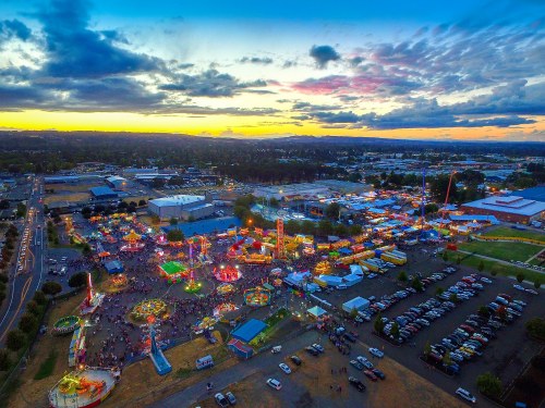 Aerial view of a vibrant fairground at sunset, with colorful lights, rides, and a bustling crowd.