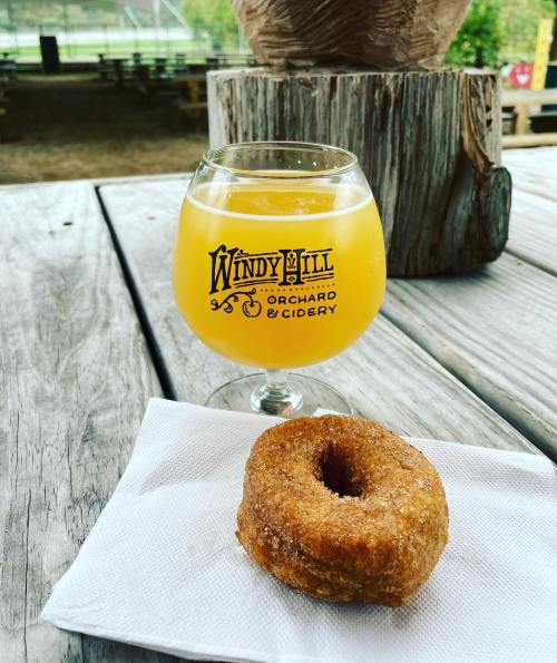 A glass of cider sits on a wooden table next to a cinnamon sugar donut on a napkin.
