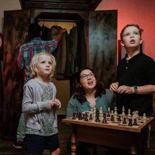 A group of three people, including two children, looks surprised while standing near a chessboard in a dimly lit room.