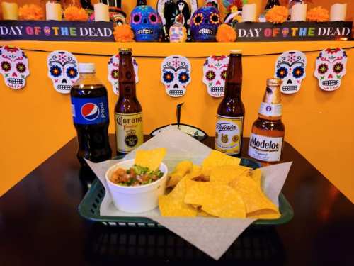 A festive table setting with chips, guacamole, and four beers, decorated for Day of the Dead with colorful skulls.