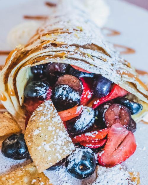 A delicious crepe filled with mixed berries, topped with powdered sugar and drizzled with sauce on a white plate.