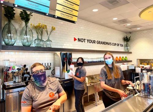 Three staff members wearing masks smile and work behind a tea counter, with colorful drinks and decor in the background.