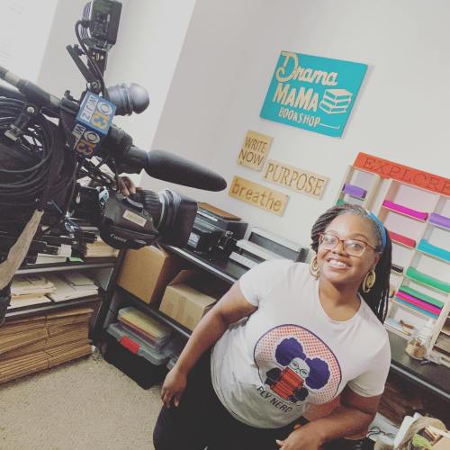 A woman in a graphic t-shirt stands next to a camera in a cozy workshop filled with colorful signs and books.
