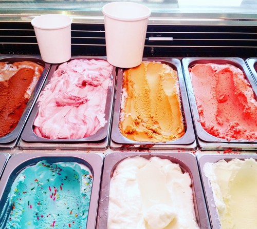 A variety of colorful gelato flavors in metal trays, with two empty cups on the side.