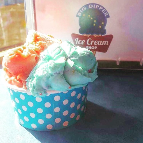 A cup of colorful ice cream with orange and mint green scoops, set against a bright background.