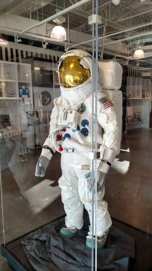 A life-sized astronaut suit displayed in a glass case, featuring a gold visor and various mission patches.