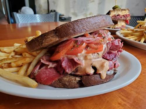 A hearty sandwich with layers of meat and tomatoes on dark bread, served with a side of fries on a wooden table.