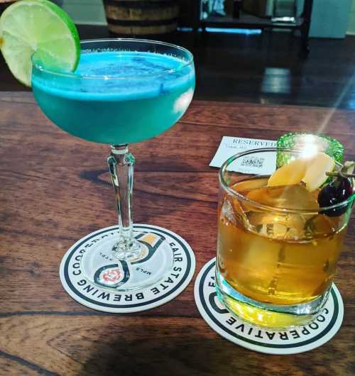 Two colorful cocktails on coasters: a blue drink with a lime slice and a brown drink garnished with a cherry and orange.