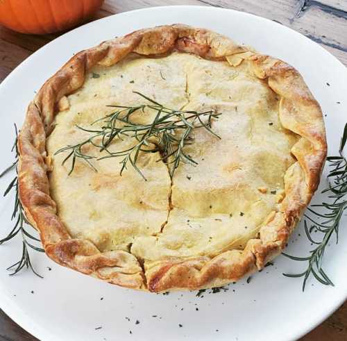 A golden-brown pie topped with herbs, resting on a white plate, with a pumpkin in the background.