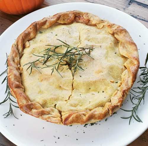 A golden-brown pie topped with herbs, resting on a white plate, with a pumpkin in the background.