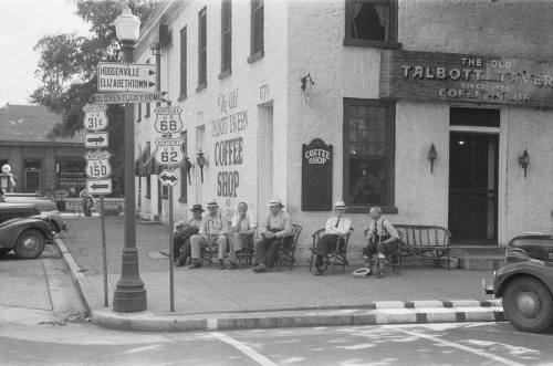 A black-and-white photo of men sitting outside a coffee shop, with vintage road signs and classic cars nearby.