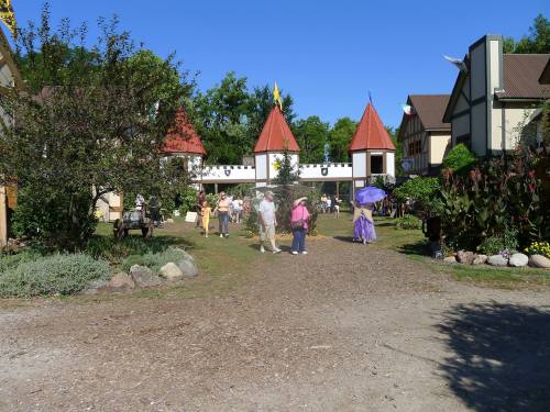 A lively outdoor scene with people walking in a fair-like setting, surrounded by trees and medieval-style buildings.