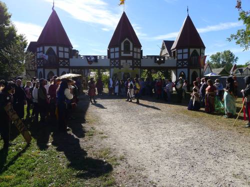 A medieval fair scene with people in costumes gathered near a castle-like entrance under a clear blue sky.