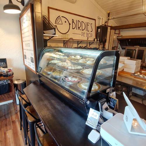 A cozy pie shop display showcasing various pies in a glass case, with a wooden counter and menu board in the background.